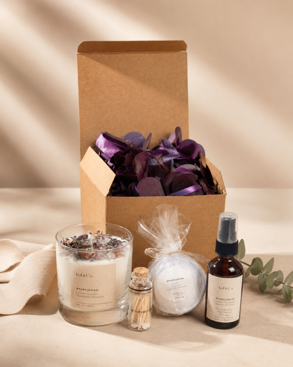 A curated spa gift set from NIKKA & KIKO on a neutral beige fabric background. The set includes a 'Resignation' candle in a glass jar, a small match bottle, a bath bomb in a clear bag, and a facial mist in a dark amber spray bottle, arranged alongside an open kraft paper box filled with purple dried flower petals.