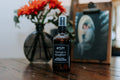 Bottle of 'Farmer's Daughter' product on a wooden surface with flowers and a photo in the background.