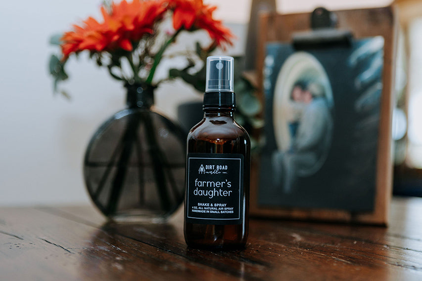Bottle of 'Farmer's Daughter' product on a wooden surface with flowers and a photo in the background.