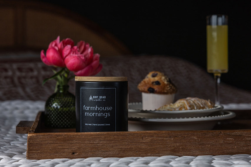 Candle labeled 'Farmhouse Mornings' on a wooden tray with flowers and pastries in the background.