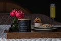 Candle labeled 'Farmhouse Mornings' on a wooden tray with flowers and pastries in the background.