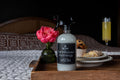 Bottle of 'Farmhouse Mornings' soap on a wooden tray with flowers, pastries, and a glass of juice.