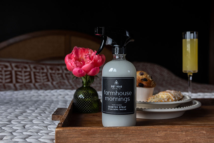 Bottle of 'Farmhouse Mornings' soap on a wooden tray with flowers, pastries, and a glass of juice.