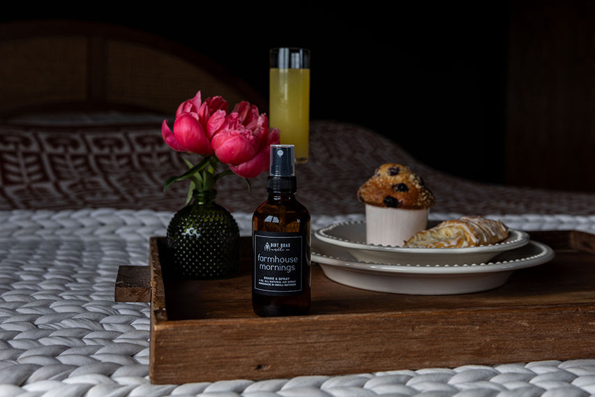 Wooden tray with a bottle of 'Farmhouse Fare' product, a vase with pink flowers, and a plate of pastries on a textured surface.