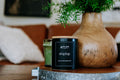 Candle labeled 'shiplap' on a wooden surface with a vase and plant in the background