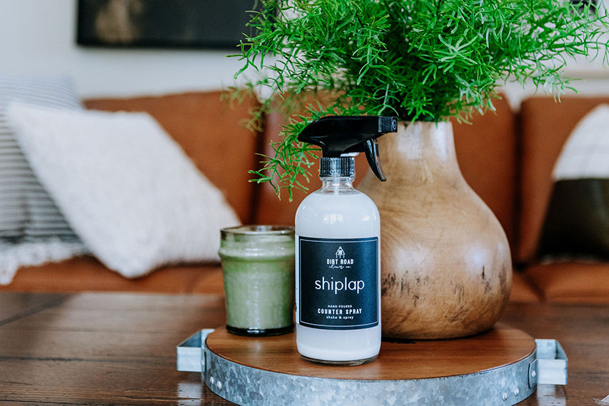 Shiplap paint can and bottle on a wooden surface with a plant and candle in the background.