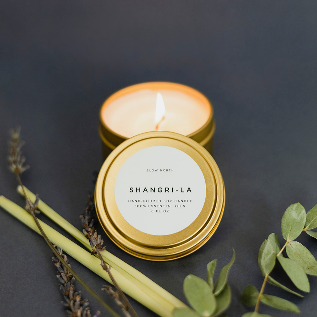Shangri-La candle with a lit wick on a dark background with greenery